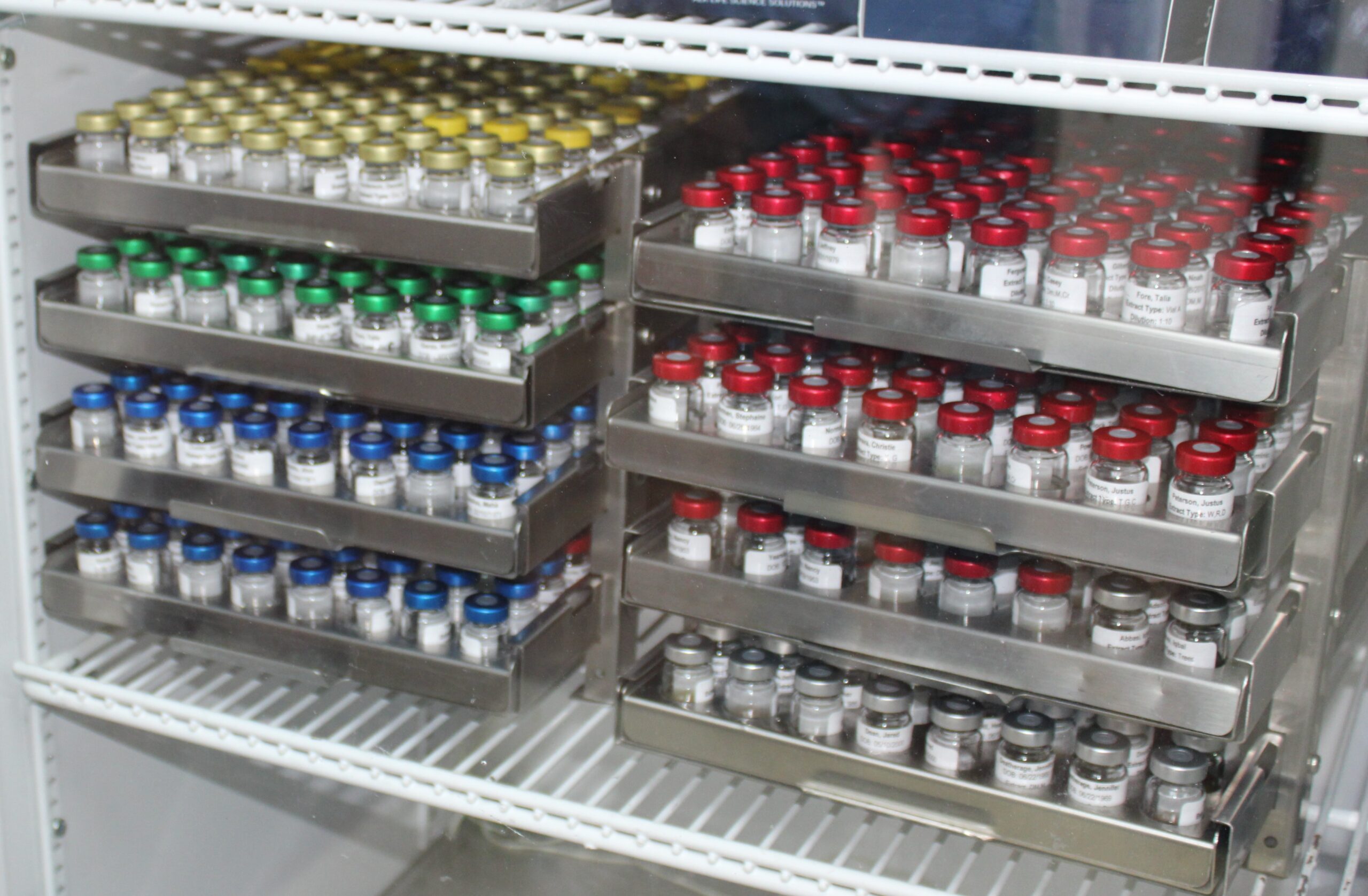 Refrigerator shelves holding metal trays of neatly organized 5 mL vials with yellow, green, blue, and red caps. Each vial labeled for pharmaceutical or lab use. Cold storage setup ensures temperature-sensitive sample integrity and inventory control.
