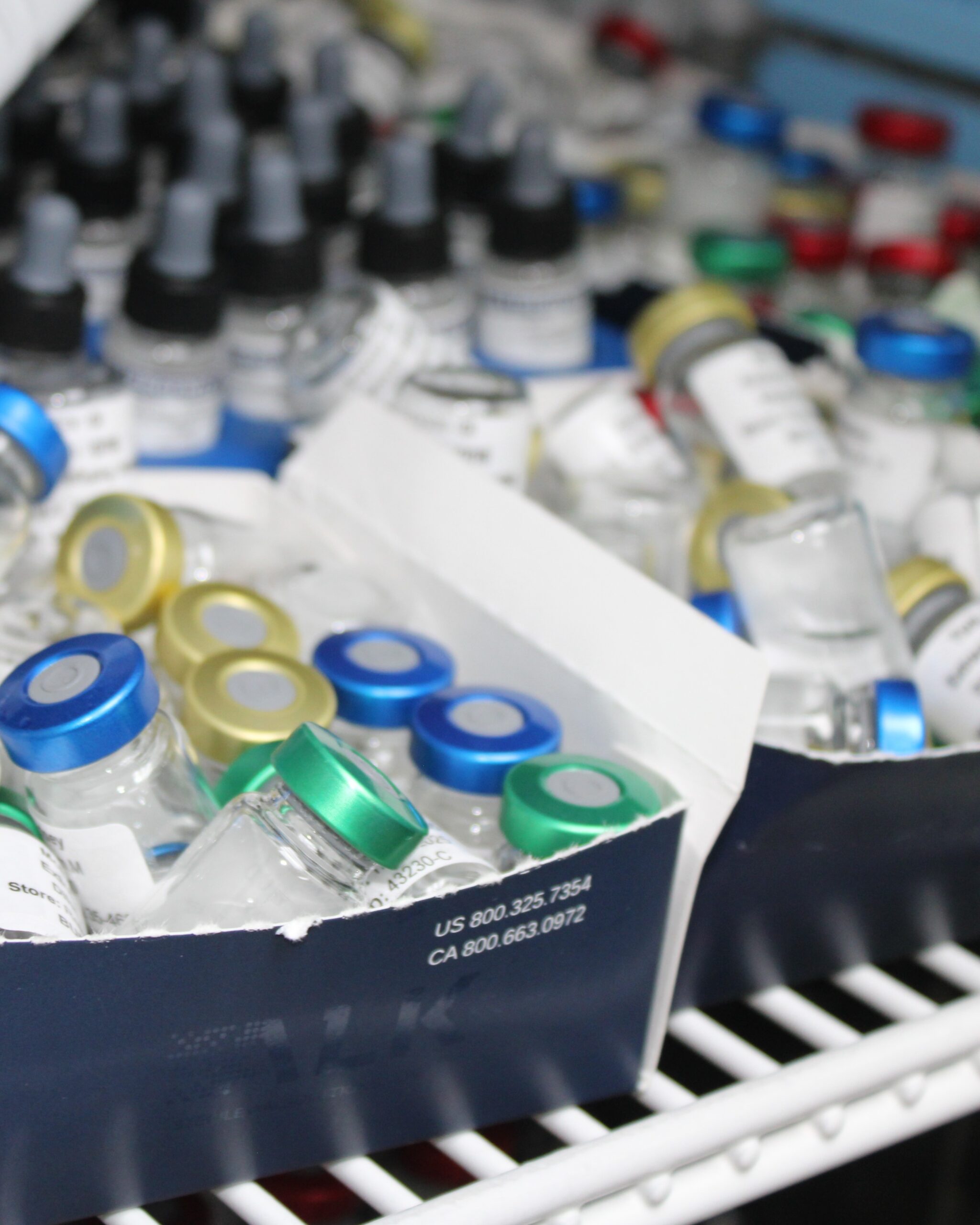 Cardboard boxes filled with glass vials featuring blue, green, gold, and red caps, stored on a white wire shelf in a medical supply area. Vials labeled for lab testing or pharmaceutical use. Contact info printed on foreground box. Background includes dropper bottles and additional vials.
